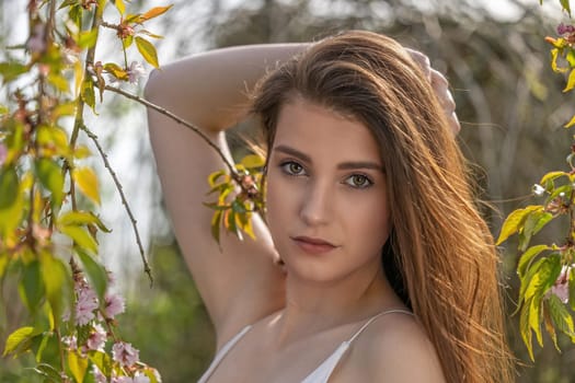 Cool brunette girl with hand in her long hair is posing on camera near sakura tree.. Horizontally.