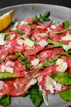 Marbled beef carpaccio with arugula and chard leaves, lemon and parmesan cheese on dark background, closeup