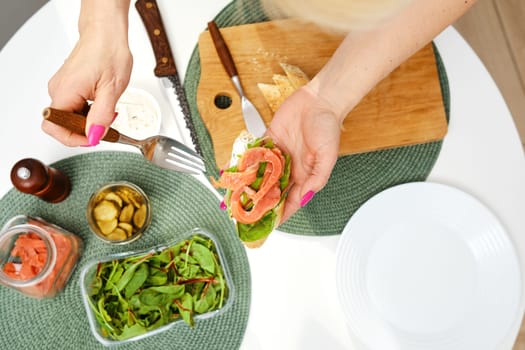 Top view of hands of middle aged woman puts smoked trout on sandwich while making breakfast