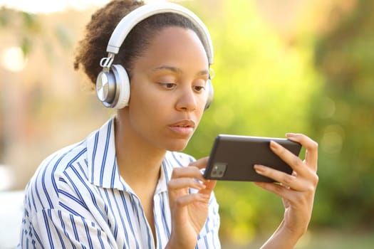 Serious black woman wearing headphone watching videos on phone in a park