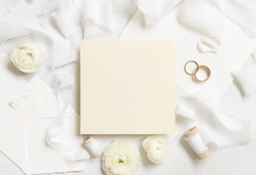 Blank card near cream roses, white silk ribbons and wedding rings top view, mockup. Romantic scene with square card and pastel flowers flat lay. Valentines, Spring or Mothers day concept