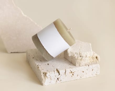 Candle with label on beige near cream travertine stones, Close up, copy space. Brand packaging mock up. Home decoration, minimal interior, Bohemian or scandinavian style