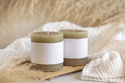 Candles with label on wooden tray near dry pampas grass and white cosy sweater Close up, copy space. Boho packaging mock up. Home decorations, Bohemian, ethnic or scandinavian style