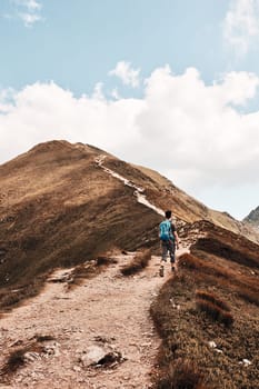 Young man with backpack hiking in a mountains, actively spending summer vacation. Rear view of teenager walking up on a hill along mountain path