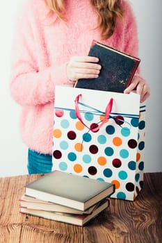 Young girl putting the books into paper bag in bookstore. A few books on a wooden table. Teenager girl wearing pink sweater and blue jeans. Vertical photo