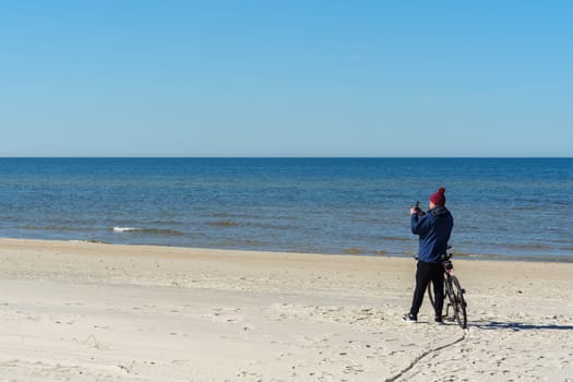 A man in warm clothes and a hat takes a photo on his phone standing near his bicycle, traveling along the Baltic Sea on the Curonian Spit, Neringa, Lithuania.
