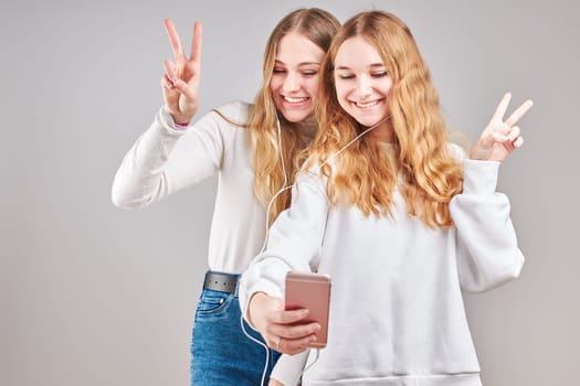 Young women girls listening to music together streaming content having fun watching video enjoying video chat talking with friends making gestures faces using smartphone earphones headphones standing over plain grey background