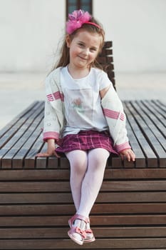 Portrait of cute adorable little girl sitting on bench in the center of town, smiling and looking at camera. Candid people, real moments, authentic situations