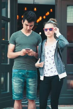 Couple of friends, teenage girl and boy,  using smartphones, talking together, standing on street in center of town, spending time together