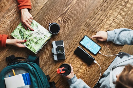 Family planning vacation trip. Girl searching travel destination and routes using navigation map on mobile phone. Woman pointing on paper map. Charging smartphone with power bank. Using technology while travelling. People sitting at wooden table with camera and backpack during journey. Travel plan concept