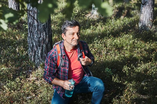 Man with backpack hiking in forest, actively spending summer vacation close to nature. Man walking on path among trees, holding bottle of water, enjoying hike on sunny vacation day