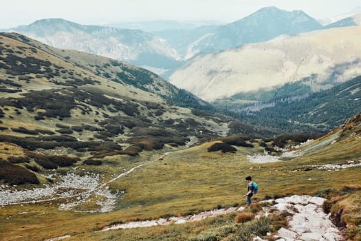 Young man with backpack hiking in a mountains, actively spending summer vacation. Rear view of teenager walking down from top of a hill to valley along mountain footpath