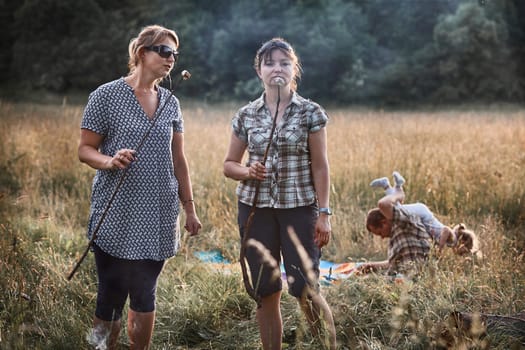 Women roasting a marshmallows over a campfire on meadow. Families spending vacations close to nature. Candid people, real moments, authentic situations