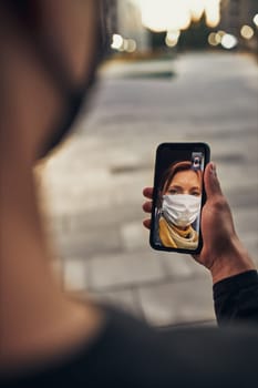 Young man having video call with her mother standing outdoors in the street in the evening wearing the face mask to avoid virus infection and to prevent the spread of disease in time of coronavirus