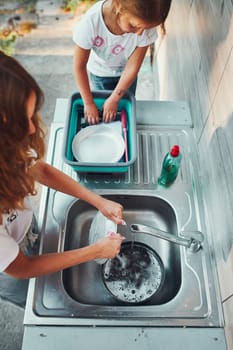 Teenager girl washing up the dishes pots and plates with help her younger sister in the outdoor kitchen during vacations on camping. Camp life. Sisters spending time and working together