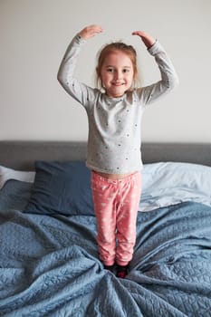 Little happy playful kid girl having fun making silly funny faces jumping on bed in bedroom in the morning at home