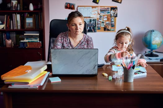 Woman mother working doing her job remotely during video chat call stream online course webinar on laptop from home while her daughter making drawings playing around and listening to stories. Woman sitting at desk in front of computer looking at screen