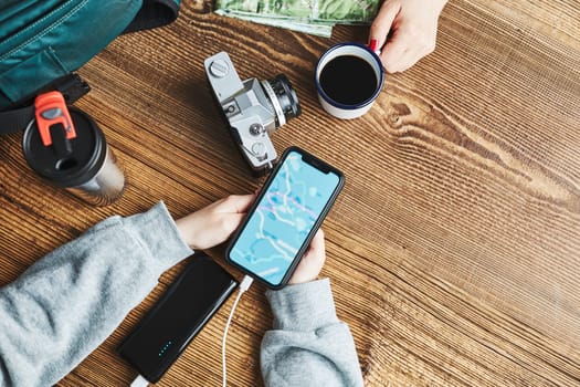 Family planning vacation trip. Girl searching travel destination and routes using navigation map on mobile phone. Charging smartphone with power bank. Using technology while travelling. People sitting at wooden table with camera and backpack during journey. Travel plan concept