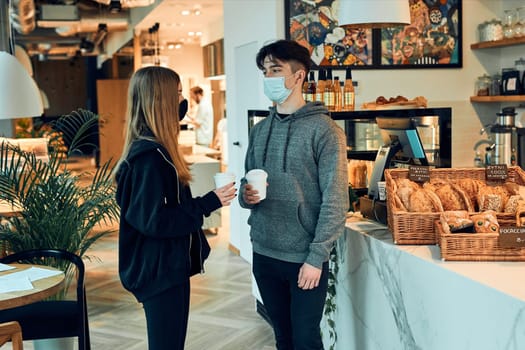 Friends having a chat, holding coffee cups, standing in a coffee shop and waiting for the orders. People wearing the face masks to avoid virus infection and to prevent the spread of disease in time of coronavirus