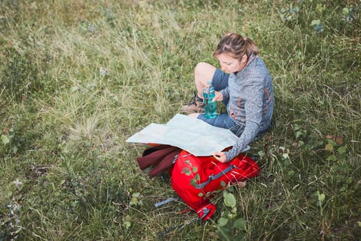 Woman with backpack having break during trip in mountains looking at map sitting on grass on summer vacation day
