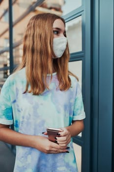 Portrait of young woman girl standing at store front downtown holding smartphone. Girl wearing face mask to avoid virus infection and to prevent the spread of disease in time of coronavirus