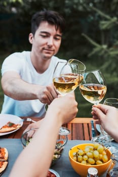Friends making toast during summer picnic outdoor dinner in a home garden. Close up of people holding wine glasses with white wine over table with pizza, salads and fruits. Dinner in a orchard in a backyard