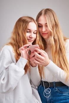 Young women girls listening to music together streaming content having fun watching video enjoying video chat talking with friends making gestures faces using smartphone earphones headphones standing over plain grey background