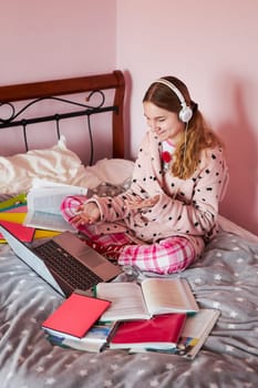 Student learning online, watching lesson remotely, listening to professor, talking with classmates on video call from home during quarantine. Young girl using laptop, headphones, books, manuals sitting in bed