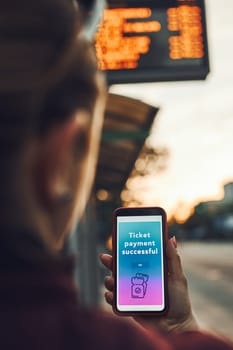 Woman buying ticket online near bus stop before ride. Person paying for ticket using mobile payment app. Using technology on the go. Order confirmation on mobile screen. Top up account in app. Bus routes schedule in background