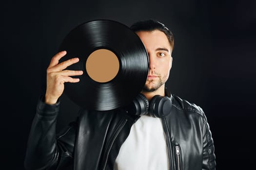 Man holding black vinyl record over face. Vintage music style. Rock style. Male wearing black jacket holding turntable disc standing on dark background. Retro music. Classic audio