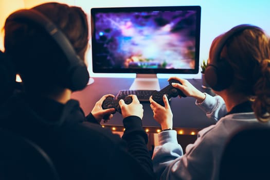 Friends playing video game at home. Gamers holding gamepads sitting at front of screen. Streamers girl and boy playing online in dark room lit by neon lights. Competition and having fun