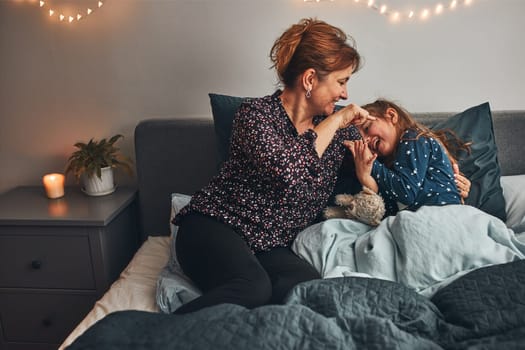Mother playing with her little daughter in bed, having fun before going sleep. Mom tickling her cute daughter before bedtime