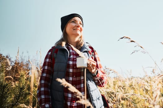 Smiling woman relaxing and enjoying the coffee during summer trip. Woman standing on trail and looking away holding cup of coffee. Woman with backpack hiking through tall grass along path in mountains. Spending summer vacation close to nature