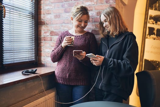 Smiling women using smartphones drinking coffee charging mobile phone relaxing in office while taking a break