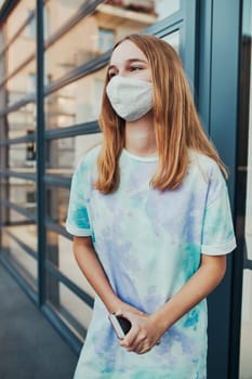 Portrait of young woman girl standing at store front downtown holding smartphone. Girl wearing face mask to avoid virus infection and to prevent the spread of disease in time of coronavirus