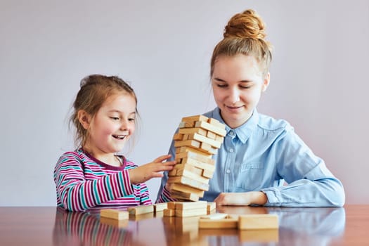 Little girl preschooler and her elder sister playing together with wooden blocks game toy. Siblings spending time sitting at desk at home