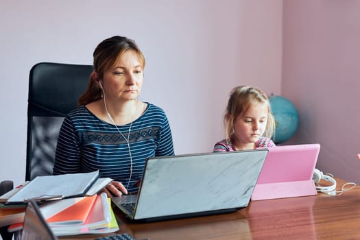 Woman mother working doing her job remotely during video chat call stream online course webinar on laptop from home while her daughter watching video on tablet. Woman sitting at desk in front of computer looking at screen