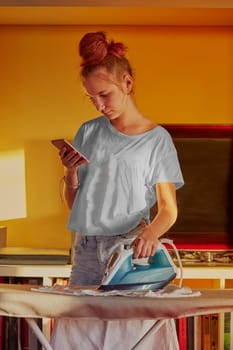 Young woman is ironing her clothes and talking on a smartphone simultaneously, standing by a ironing board in a room at home. Routine housekeeping task at home. Candid people, real moments, authentic situations