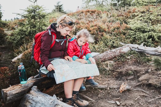 Family trip in mountains. Mother and her little daughter examining a map, sitting on stump, taking break during summer trip