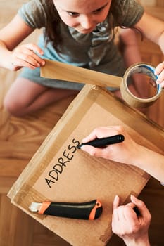 Woman writing an address on a cardboard box parcel in room at home. Little girl helping her mother to wrap a package. Preparing parcel to send