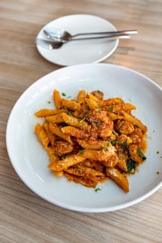 Spicy Italian Sausage Penne In a round white plate, next to it is an empty plate with cutlery, on a wood grain table, natural light. Selective focus.
