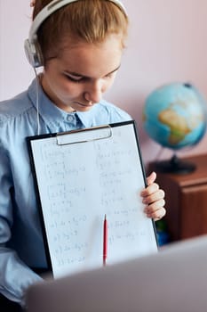 Young woman student showing homework, having classes, learning online, watching lesson remotely, listening to professor, talking with classmates on video call from home during quarantine. Young girl using laptop, headphones, books, manuals sitting at a desk
