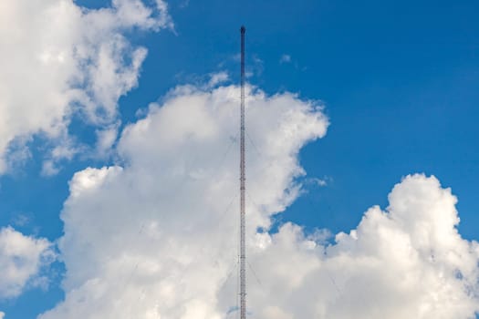 A radio antenna made of a steel frame sits in the center of the picture with a sling cable fixed to the ground. The background is a bright blue sky with white clouds.