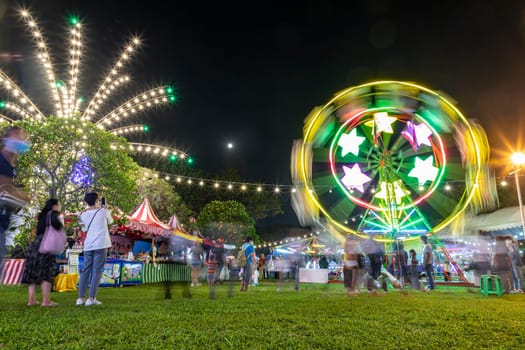 Bangkok, Thailand - Dec 8, 2022 : People take a walk near the spinning Ferris wheel at the Red Cross Fair at Lumpini Park from December 8-18, 2022. The picture is blurred by movement.