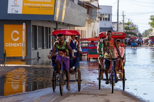 Toliara, Madagascar - November 21. 2022: Traditional rickshaw bicycle with Malagasy passengers on the street, one of the ways to earn money. Everyday life on the cities street of Madagascar.