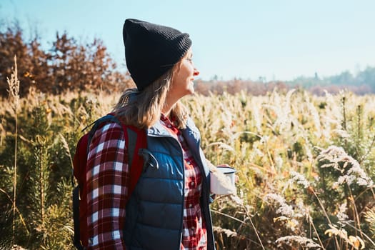 Woman taking break and enjoying the coffee during vacation trip. Woman standing on trail and looking at view. Woman with backpack hiking through tall grass along path in mountains. Spending summer vacation close to nature