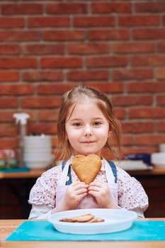 Little girl getting happy out of baked cookie made oneself. Kid taking part in baking workshop. Baking classes for children, aspiring little chefs. Learning to cook. Combining and stirring prepared ingredients. Real people, authentic situations