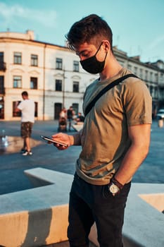 Young man standing in the city center in the evening, using smartphone, wearing the face mask to avoid virus infection and to prevent the spread of disease in time of coronavirus