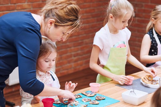 Little girl with her mom's help decorating baked cookies with colorful sprinkle and icing sugar. Kid taking part in baking workshop. Baking classes for children, aspiring little chefs. Learning to cook. Combining and stirring prepared ingredients. Real people, authentic situations
