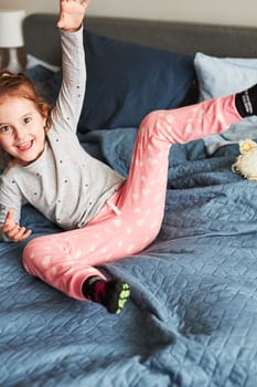 Little happy playful kid girl having fun making silly funny faces jumping on bed in bedroom in the morning at home